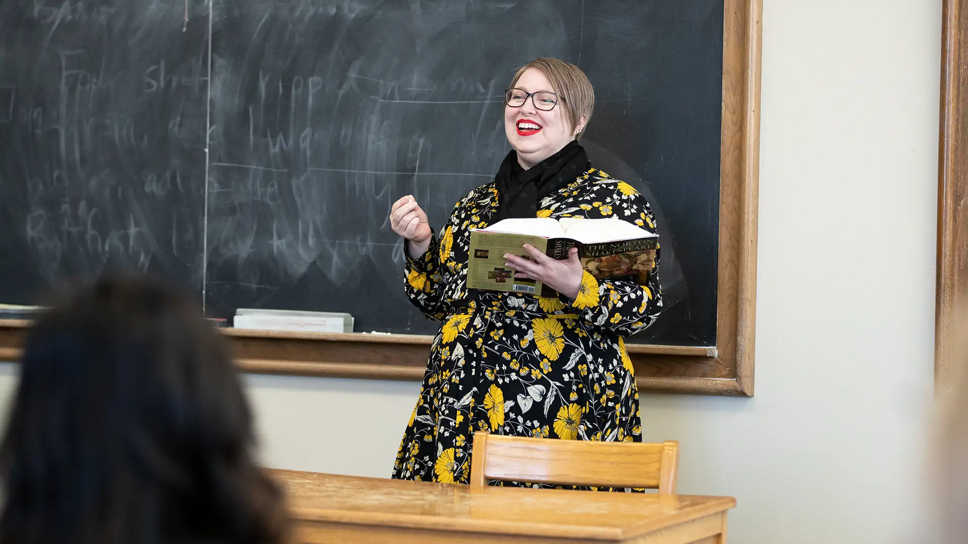 Rachel Clark stands in front of a blackboard with an open book in her hand.