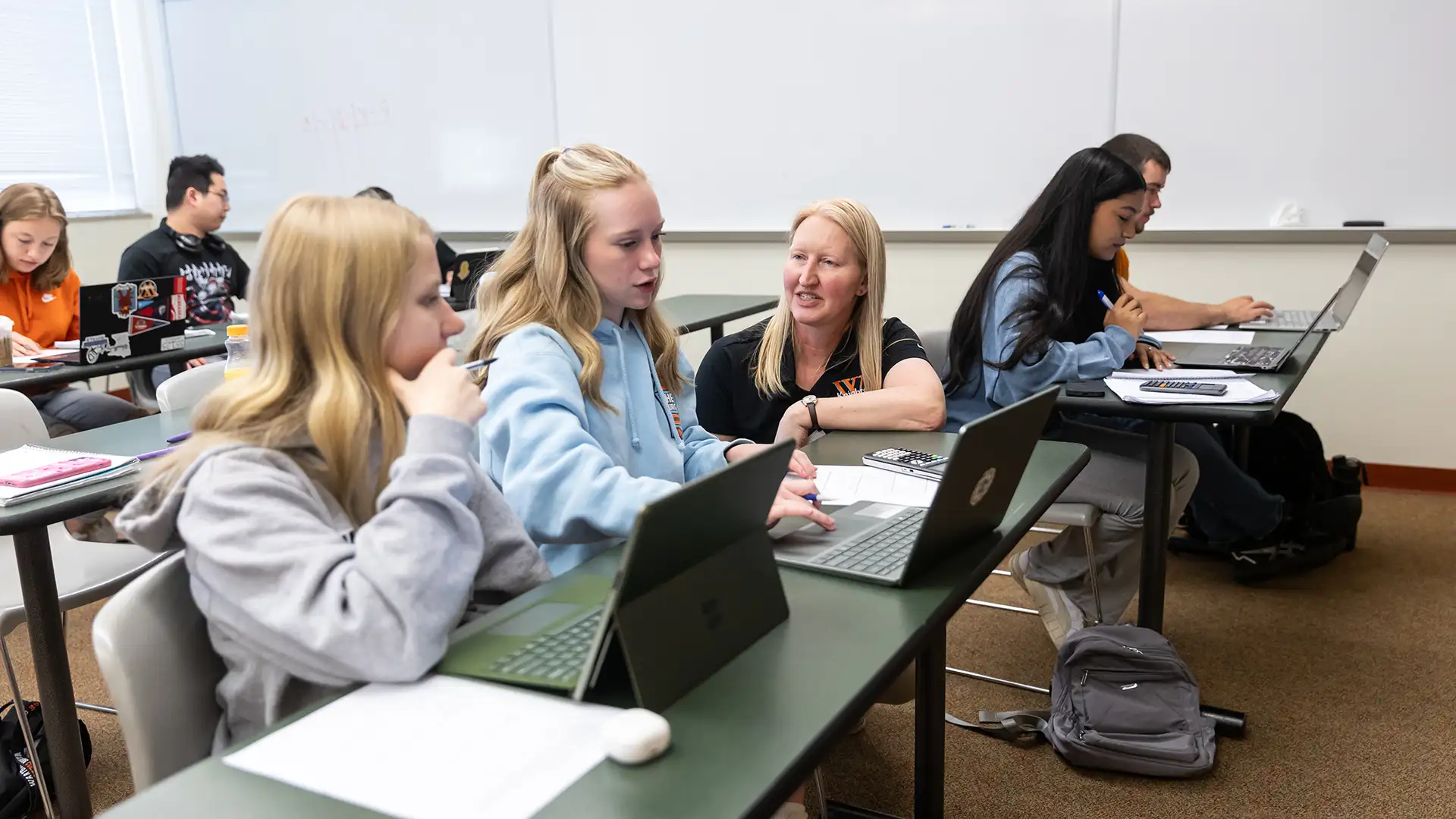 Professor kneels down to help two female students with computers open in front of them on the table during a math class. 