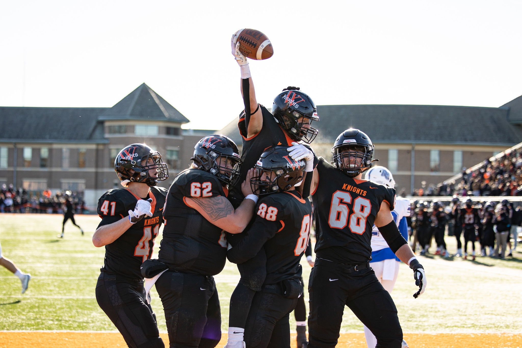 Football players celebrating a touchdown. One is hoisting the football over the group.