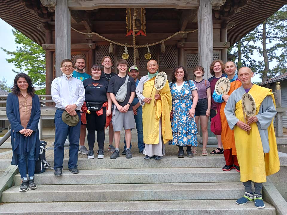 Students stand on the steps outside a religious building in Japan with individuals in traditional religious dress