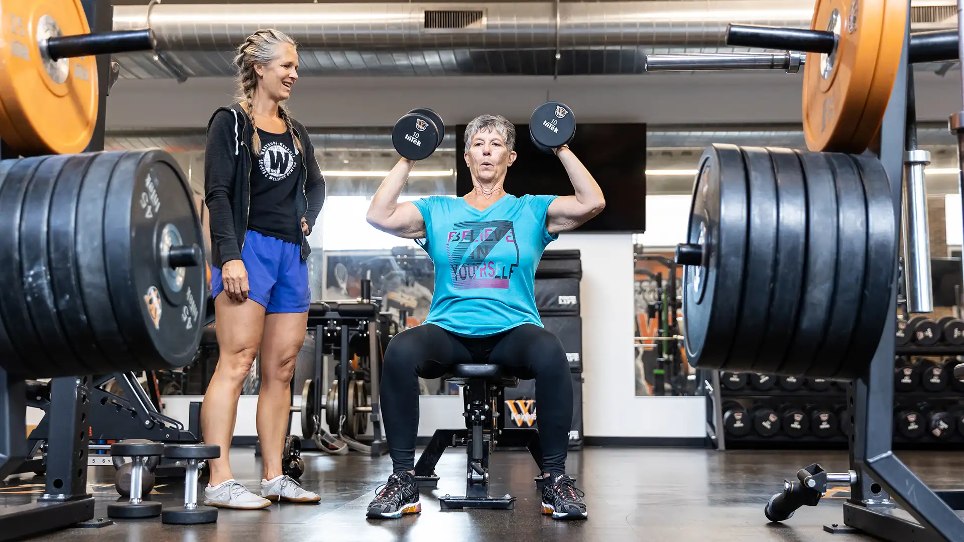 Female personal trainer helps female client who is seated and attempting to lift two weights over her head.