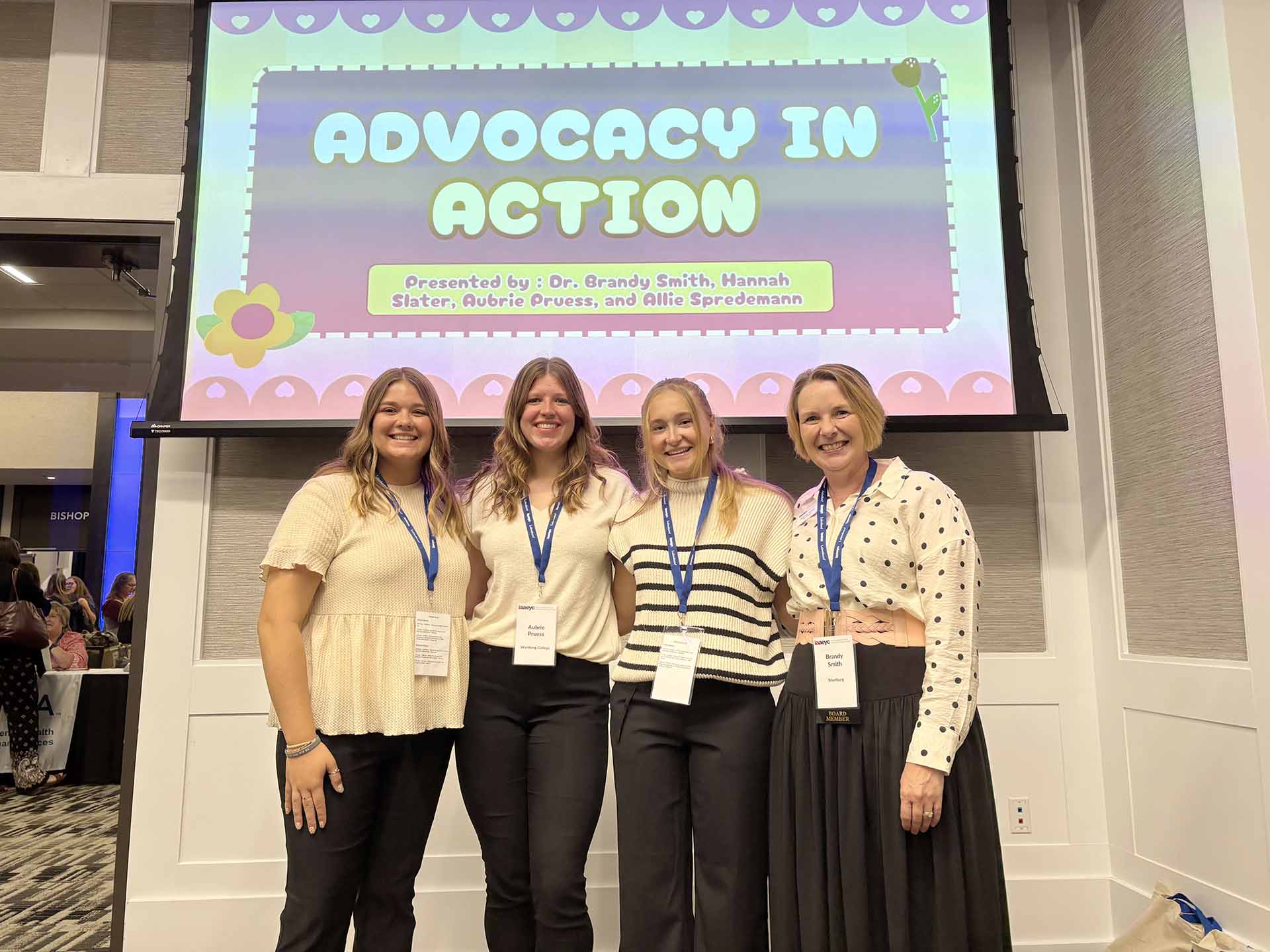 Three students and a professor stand in front of a project screen with their presentation title slide on the screen.
