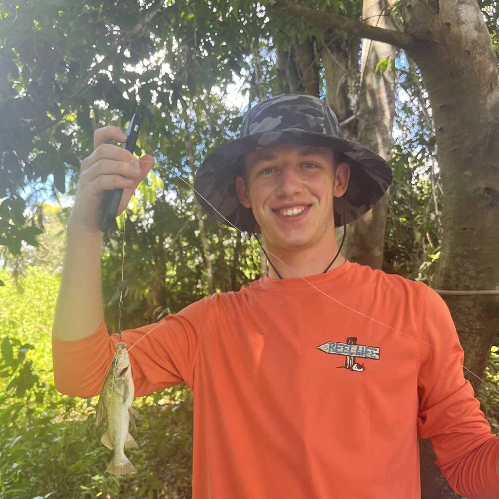 Colton Abels wears a fishing hat and bright orange shirt. He is holding a small fish on a string. 