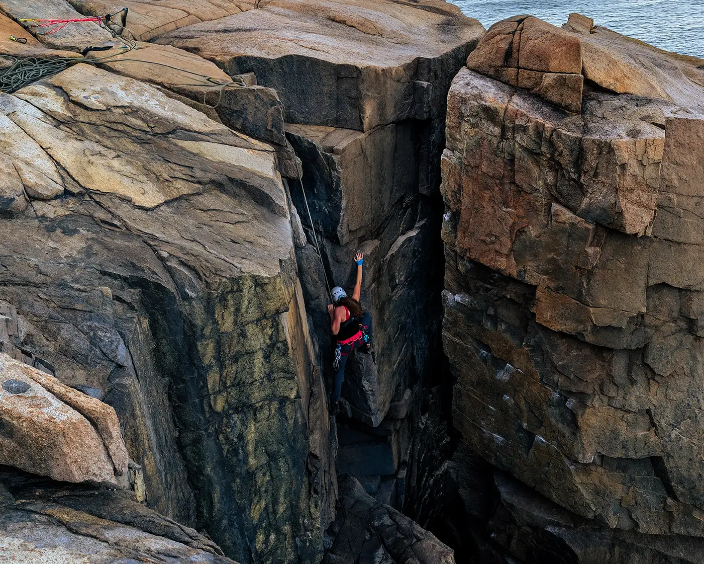 Natalie Bonthius scales a large cliff front in Acadia National Park. Water is in the background.