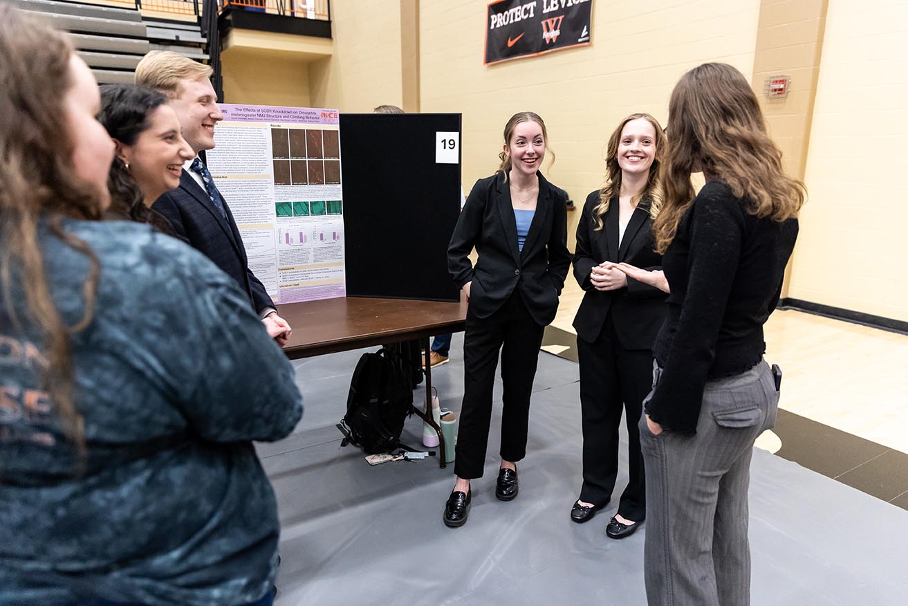 Two female students stand in front of a table with a research poster board. They are presenting their findings to a small group of people in Levick Arena during RICE Day.