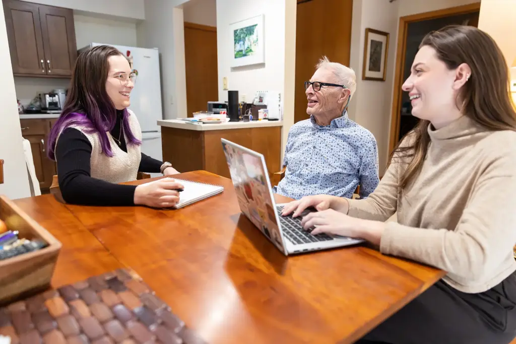 Alexa Brockmueller, Dale Johnson, and Mya Baker sit around a small dining room table. Alexa has a notebook in front of her while Mya has her computer open and hands ready to type. All three are smiling. 