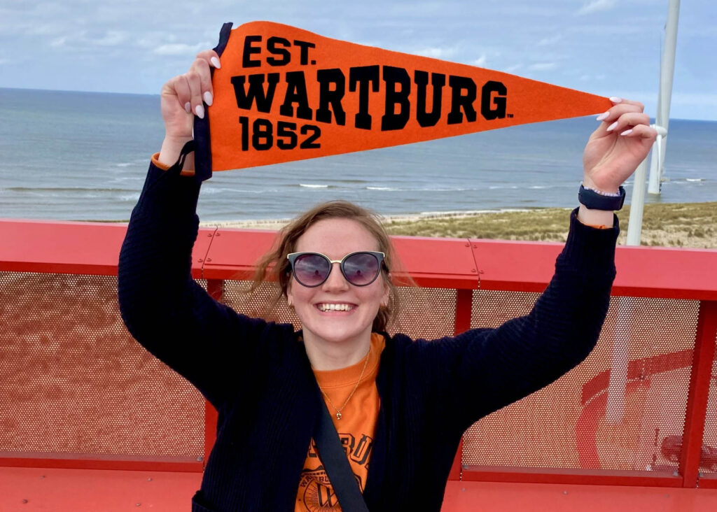 Brandy Beatty holds a Wartburg pennant over her head with the beach and water in the background. 