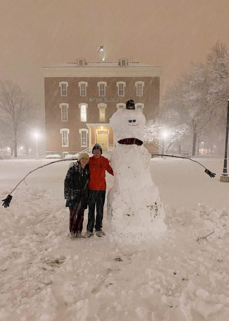 Brandy Beatty and her boyfriend stand beside the snowman they built. It is snowing and Old Main is in the background