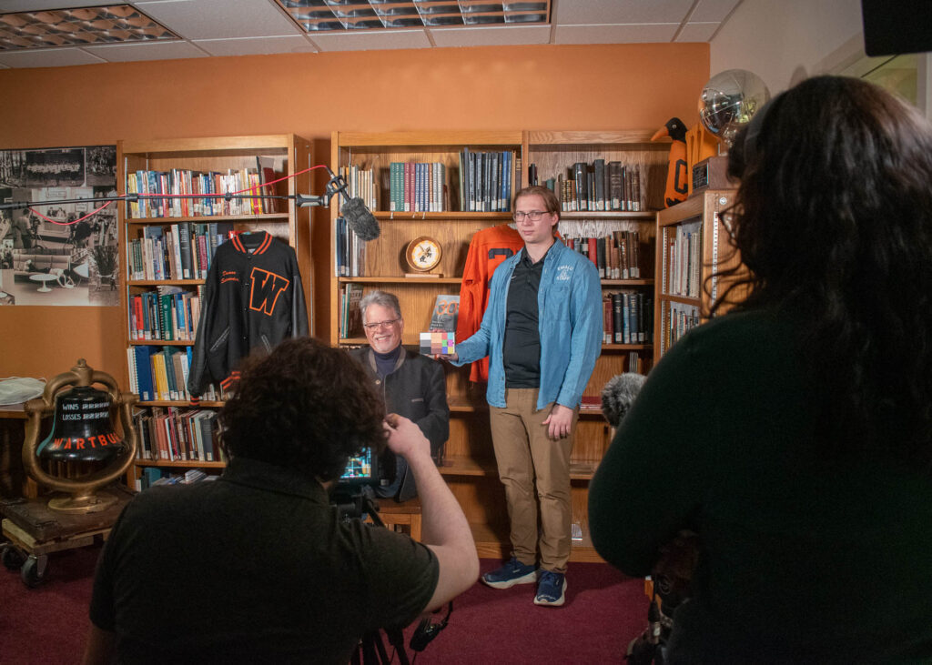 Caleb Kammerer stands with Randall Schroeder (seated) in the Wartburg College Archives while other members of his team test the camera. Schroeder worked in Vogel Library from 1995 to 2007, and his father, Duane, was the college's sports information director for 42 years.