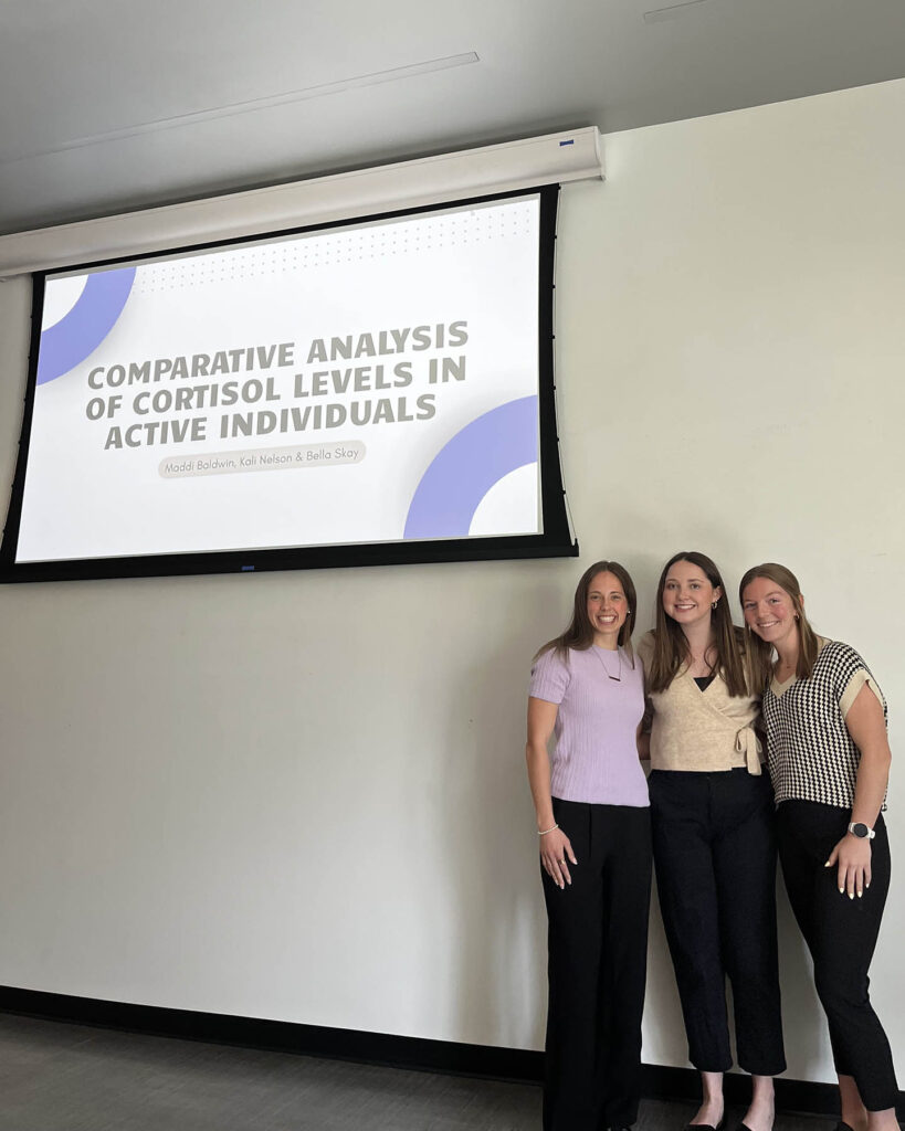 Kali Nelson stands with her research group in front of a projector screen that reads Comparative Analysis of Cortisol Levels in Active Individuals.