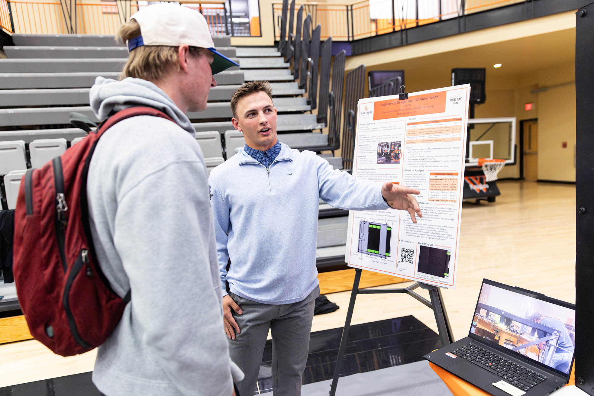 Aiden Tobin stands in front of his poster at RICE Day explaining his project to a fellow student