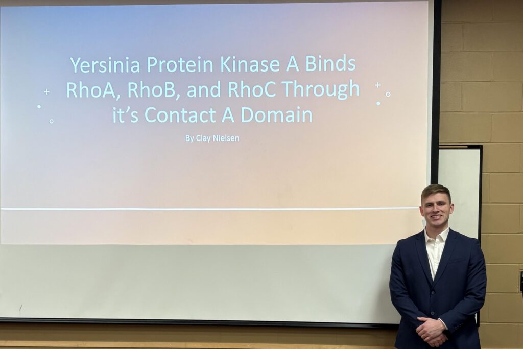 Clay Nielsen stands behind a screen displaying the title of his research, Yersinia Protein Kinase A Binds RhoA, RhoB, and RhoC through it's contact A Domain.