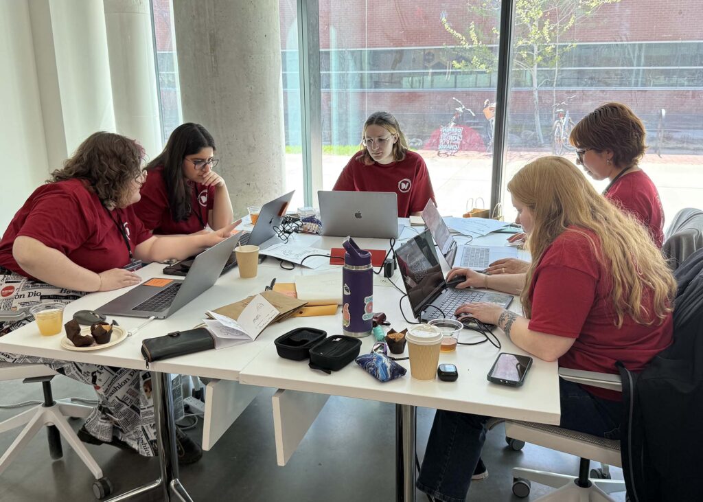 Five students sit around a square table with computers in front of them. 
