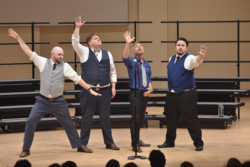 The Newfangled Four on a stage with risers behind them. The men are singing and holding their arms up in an expressive manner.