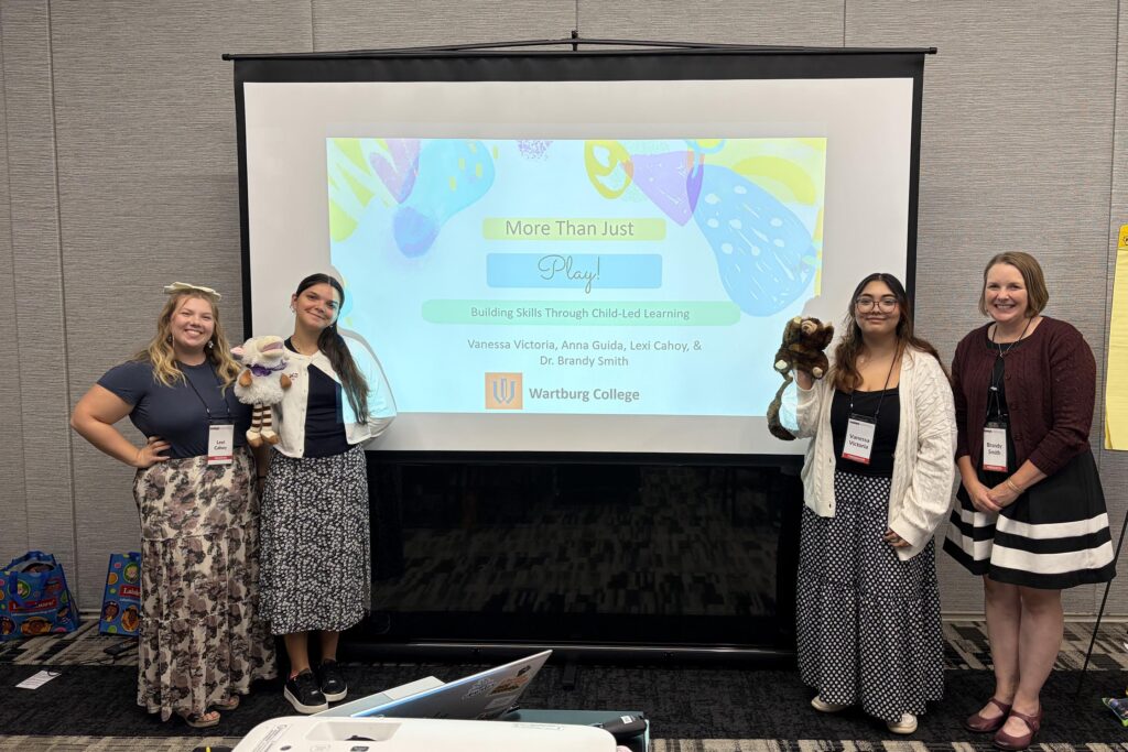 From left Lexi Cahoy, Anna Guida, Vanessa Victoria, and Brandy Smith stand beside a screen projecting the title of their presentation.