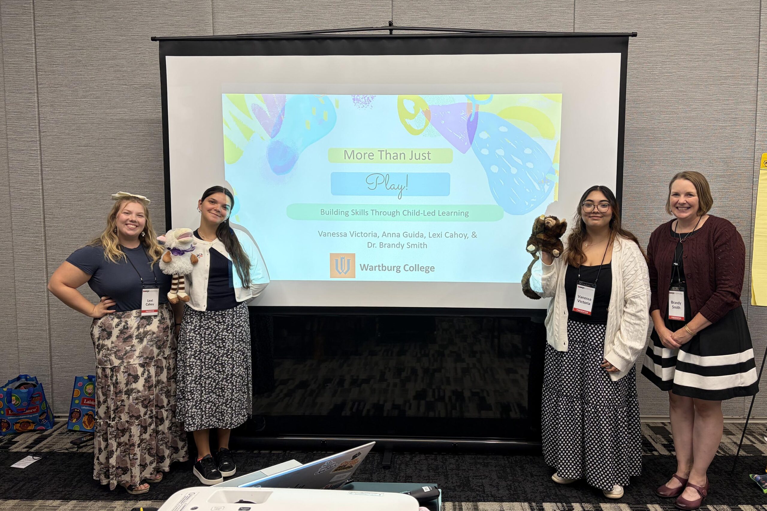 From left Lexi Cahoy, Anna Guida, Vanessa Victoria, and Brandy Smith stand beside a screen projecting the title of their presentation.